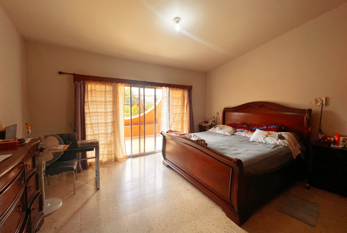 Habitación principal para con una cama tamaño Queen, cómoda de madera, al lado un escritorio de trabajo y una ventana corrediza con acceso al balcón con vista a la Res. Centroamérica.