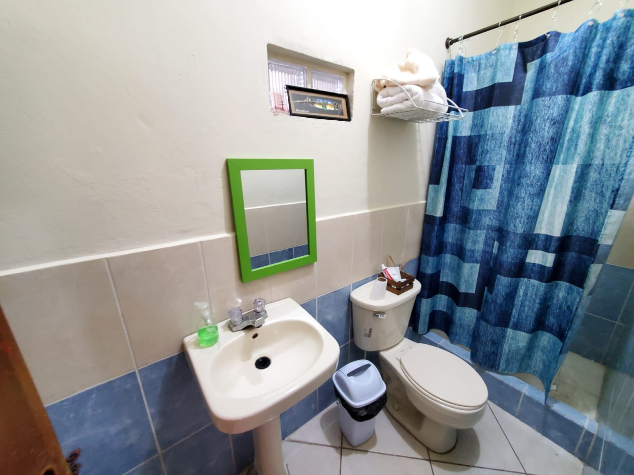 Baño con su inodoro, lavamanos, espejo, una estante para poner toallas y una cortina de baño azul en la colonia Modelo, Honduras.