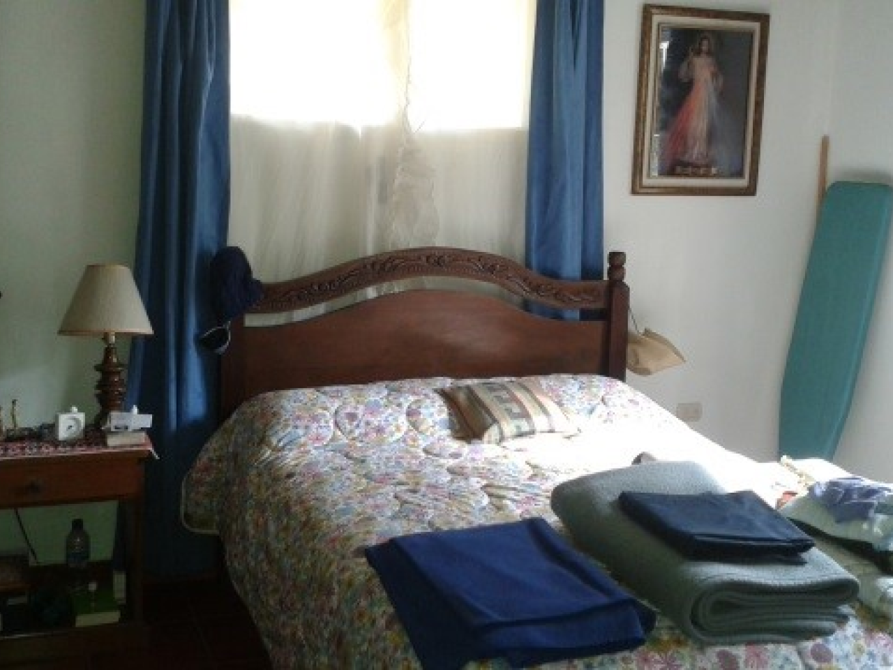 Habitación principal con una cama matrimonial bese de madera junto una mesita de noche color cafe y una ventana con cortina de color blanco.