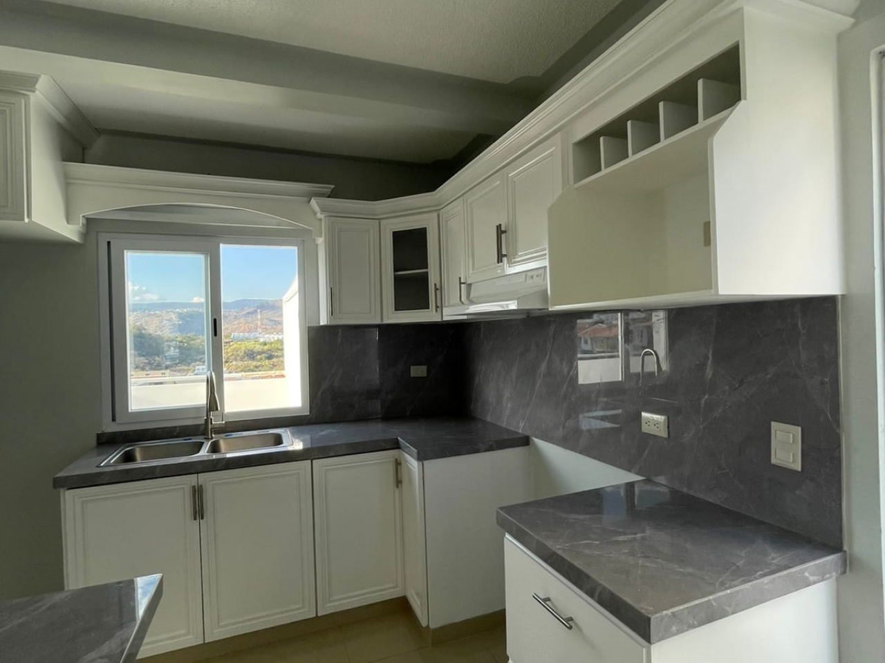 Cocina amplia con muebles de madera color blanco, plancha granito color gris y una ventana con vista a la residencial.