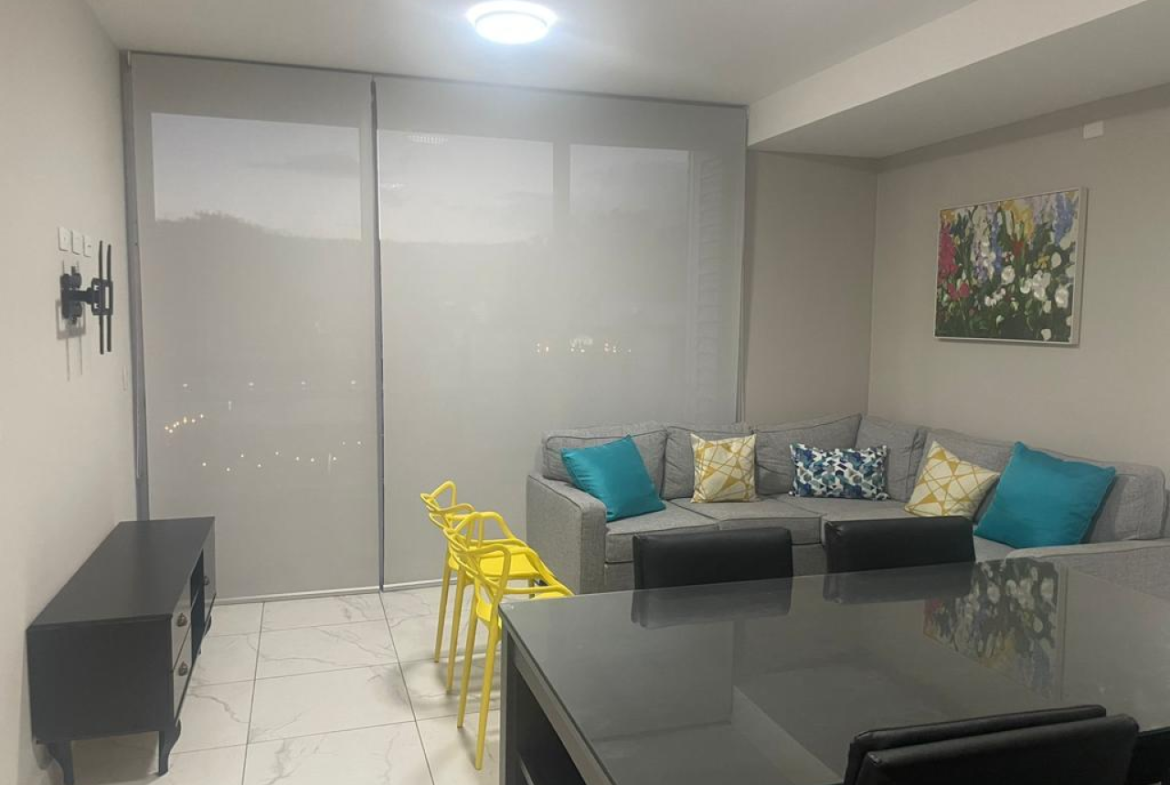 Sala de estar con piso cerámica color blanco, paredes color beide, sofá tipo L, cuadro de pintura, comedor negro y una puerta corrediza con cortinas roller color gris.