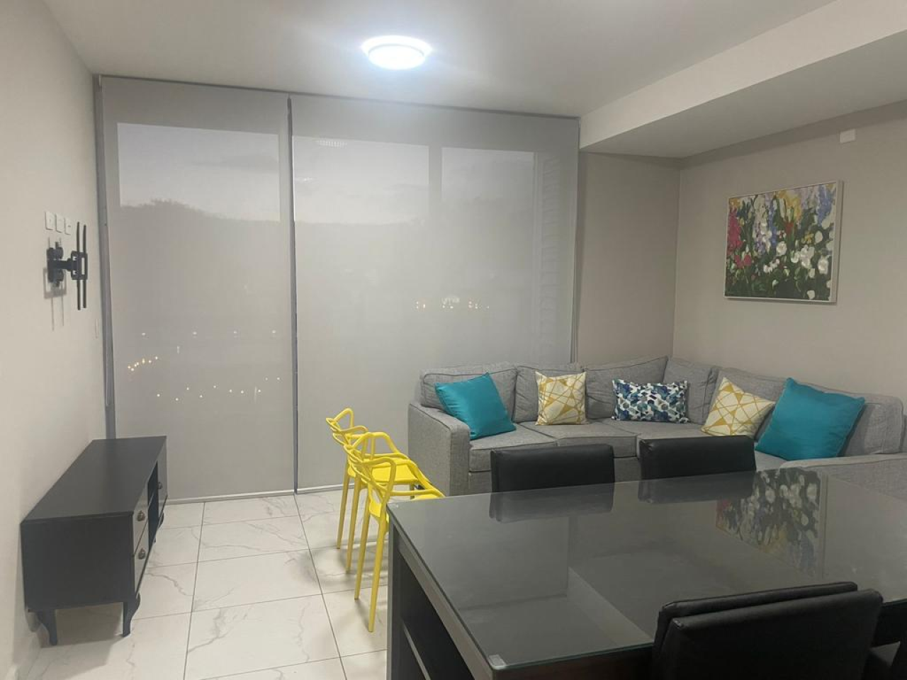 Sala de estar con piso cerámica color blanco, paredes color beide, sofá tipo L, cuadro de pintura, comedor negro y una puerta corrediza con cortinas roller color gris.