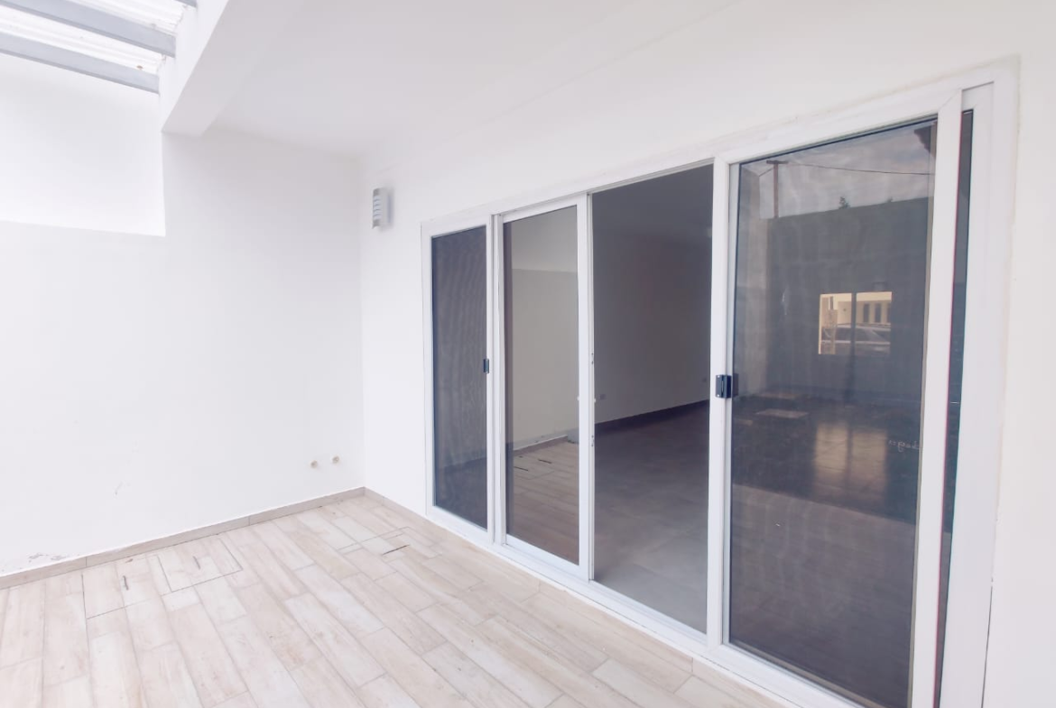 Puerta de vidrio corrediza con acceso a la sala y patio trasero, pared de concreto color blanco y piso de cerámica estilo madera.