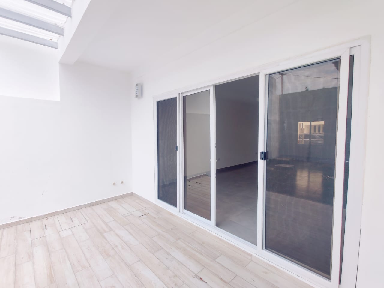 Puerta de vidrio corrediza con acceso a la sala y patio trasero, pared de concreto color blanco y piso de cerámica estilo madera.