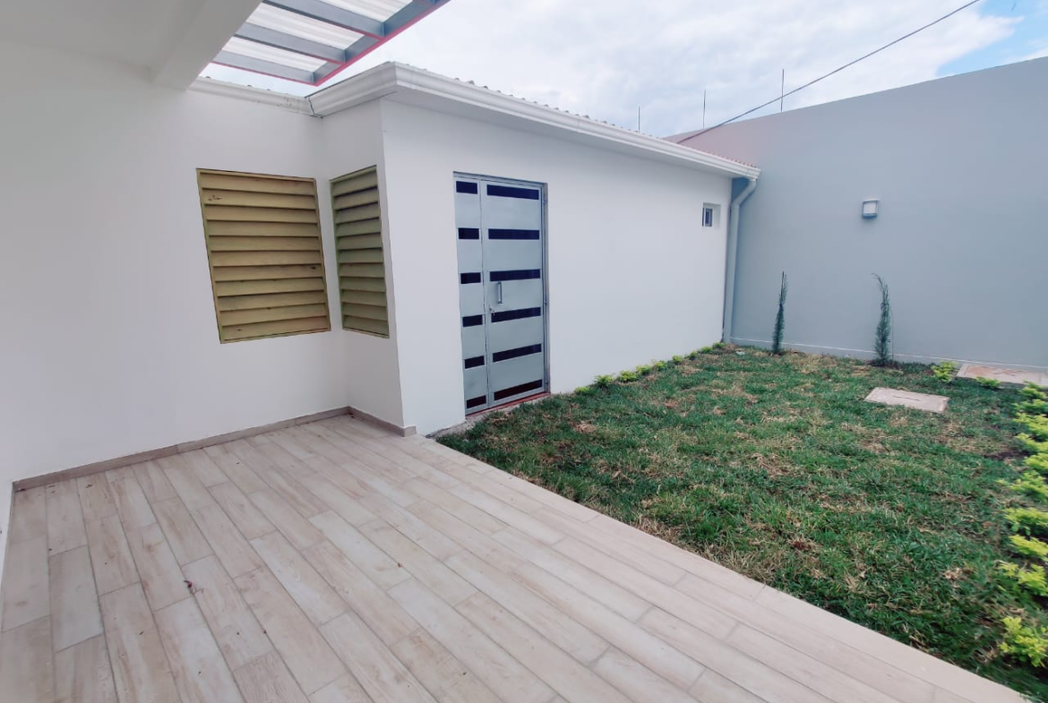 Patio trasero, pared de concreto color blanco y piso de cerámica estilo madera, junto a bodega y patio con césped.
