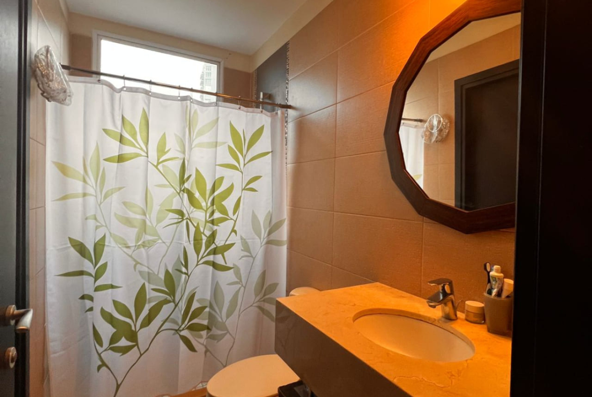 Baño con regadera moderna color gris oscuro, retrete y lavado de cerámica con espejo redondo con bordes de madera y luces modernas que dan ambiente cálido y acogedor.