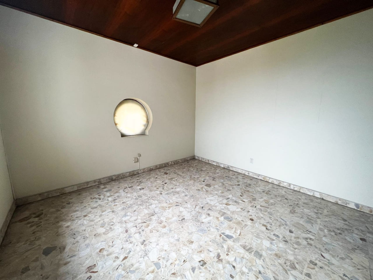 Amplia habitación con paredes color blanco, techo de madera con lámpara colgante y una ventana redonda con vista al jardín.