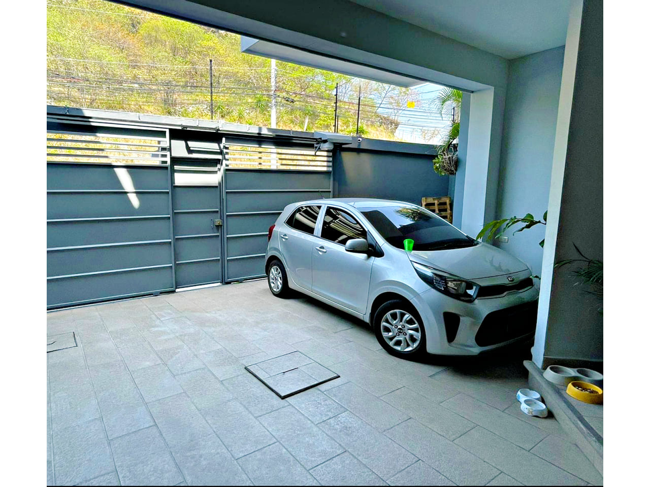 Amplio garaje para dos vehículos, ubicado en la parte frontal de la casa, con portones de color gris un poco oscuros pero que dan la sensación de entrar a una casa moderna.