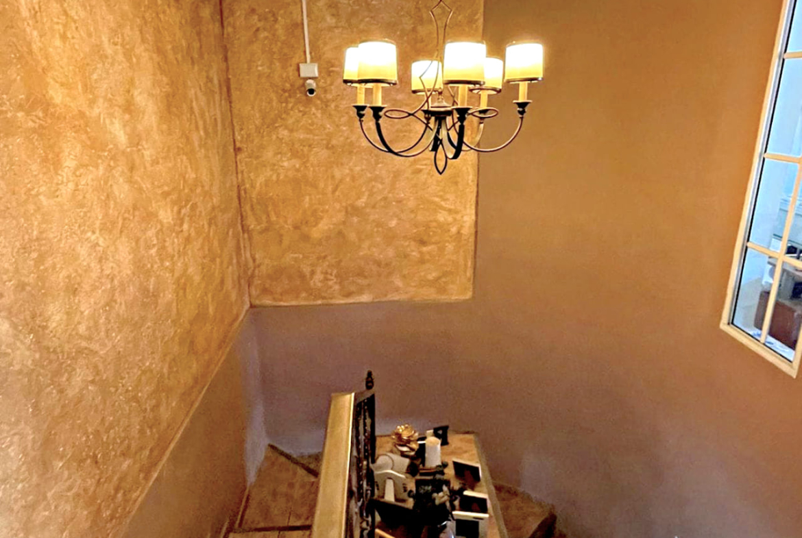 Escaleras que brindan acceso al segundo piso, con un candelabro que ilumina bellamente la zona, con una pequeña ventana al exterior.