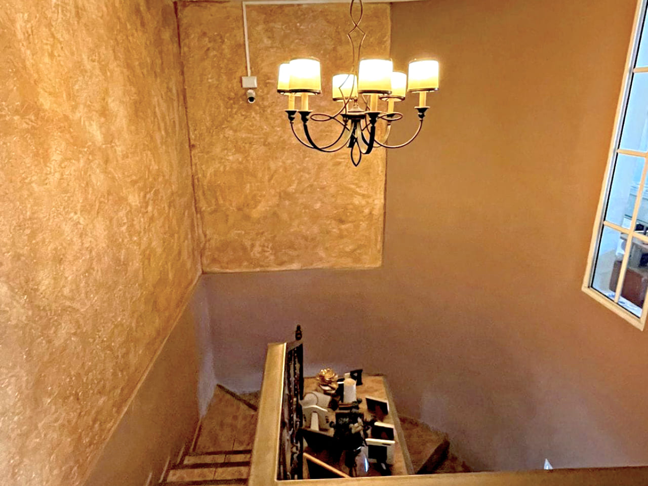 Escaleras que brindan acceso al segundo piso, con un candelabro que ilumina bellamente la zona, con una pequeña ventana al exterior.
