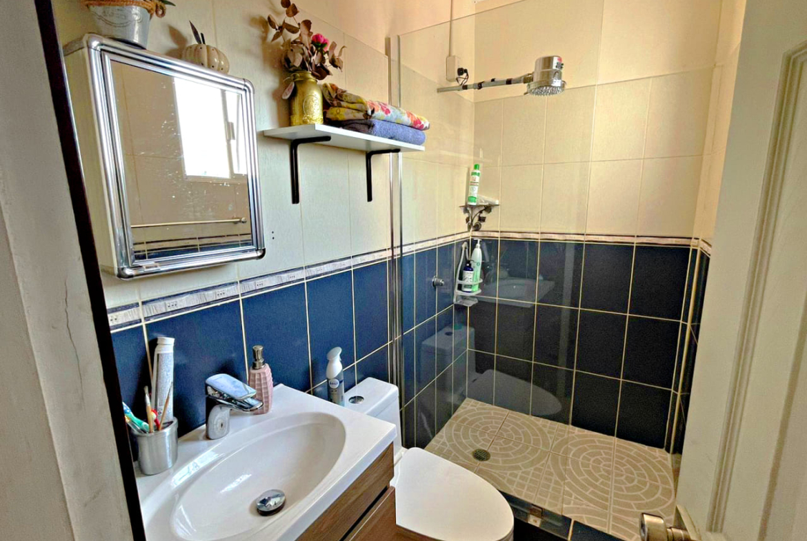 baño con regadera y suelo de cerámica con división de vidrio, un inodoro color blanco y una lavamanos de cerámica con un pequeño espejo de puesta.