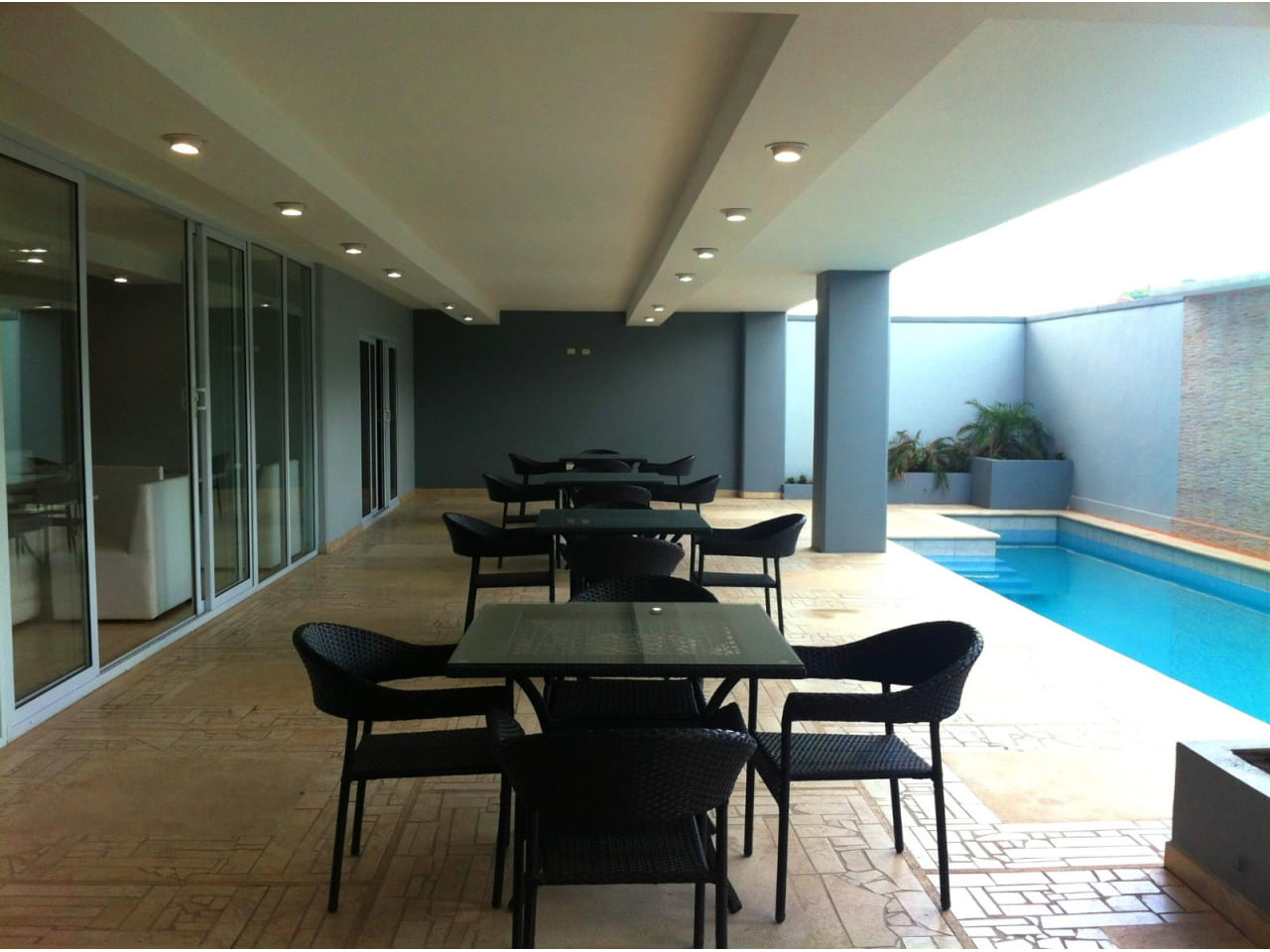 Área de piscina con diferentes mesas y sillas para disfrutar del ambiente refrescante de la piscina., paredes de color gris oscuro, suelo antideslizante de color café claro.
