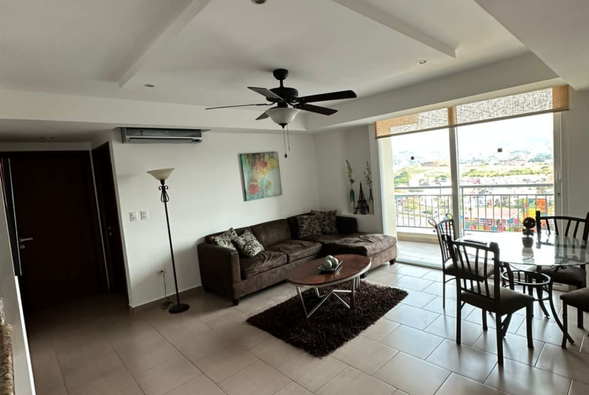 Sala de estar con televisor, con sofas de color café oscuro, lampara elegante de pie, recuadros abstractos colgados en la pared, acceso al área de balcón.