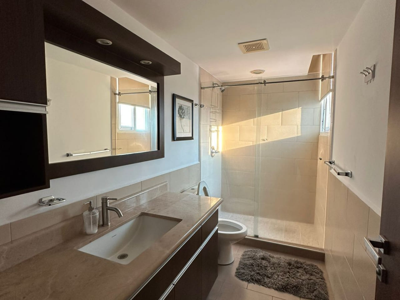 Baño completo con paredes de color blanco, regadera de cerámica, con retrete de color blanco, lavamanos moderno con mesa con superficie de cerámica, amplio espejo.