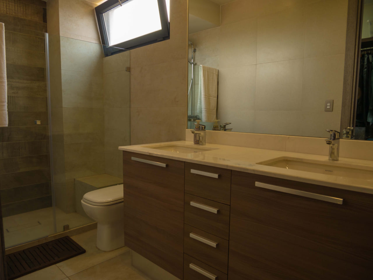 Baño con un mueble de madera, plancha de granito, con un espejo grande junto a ducha con puerta de vidrio.