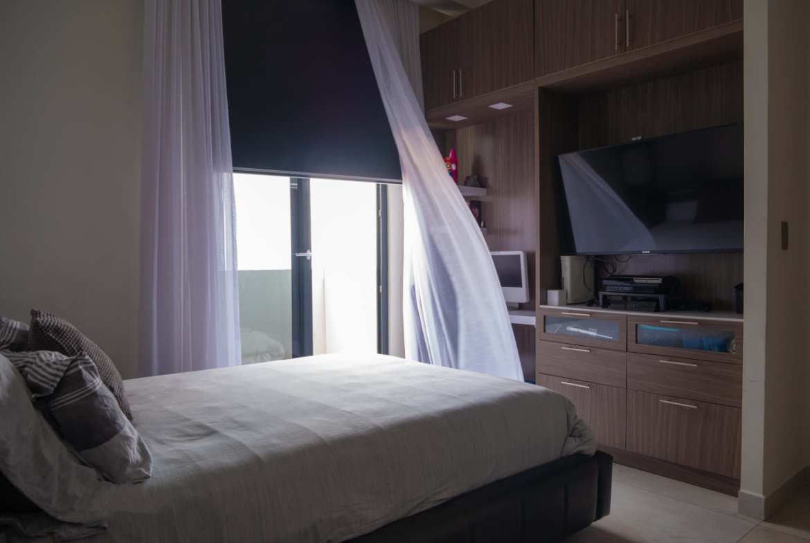 Habitación principal con una cama queen, en frente un televisor plasma, debajo una comoda y junto a él una ventana grande con vista de la ciudad de Tegucigalpa.