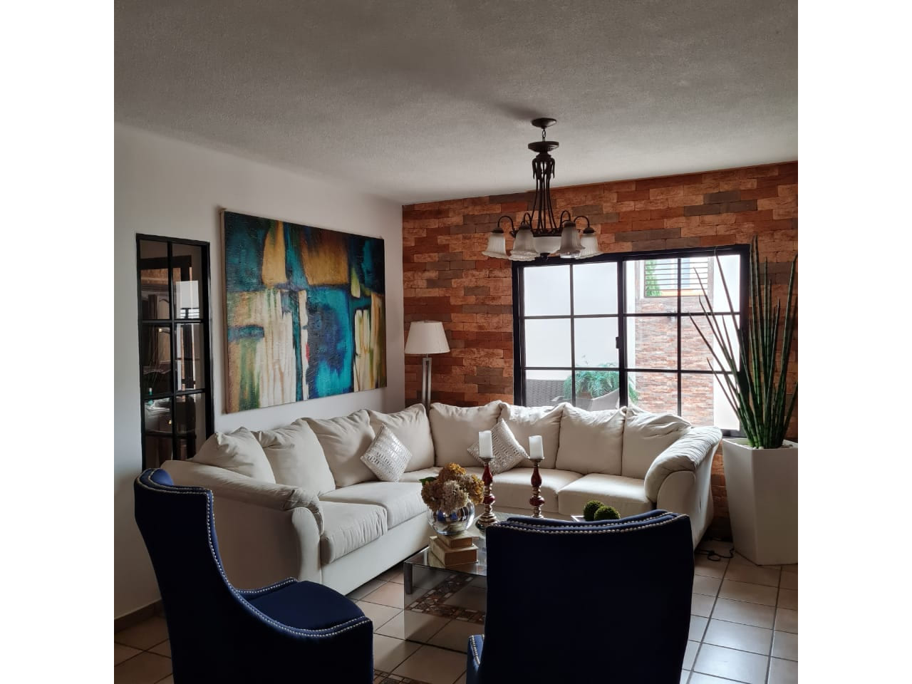 Sala de estar con muebles de color blanco, una lampara grande colgante, un cuadro grante detras junto a una puerta corrediza con acceso al patio.