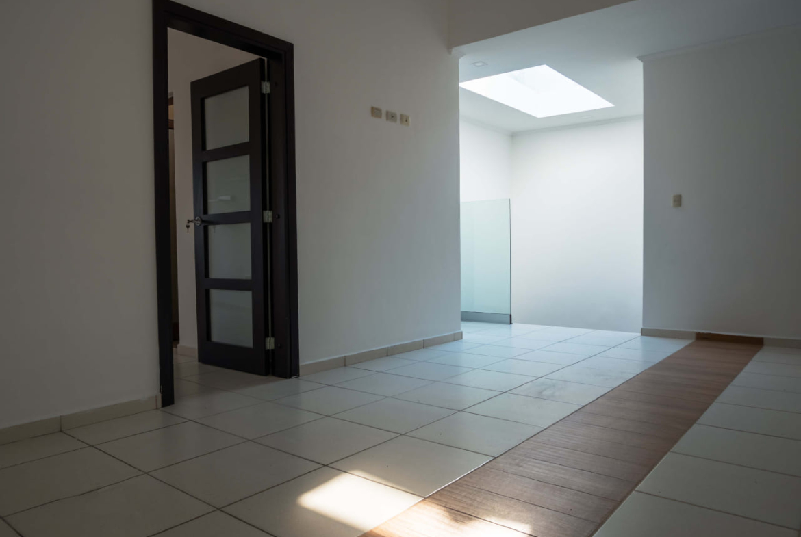 Paredes de color blanco con suelos de cerámica clara, dan acceso a diferentes habitaciones que cuentan con puertas de madera oscura adornadas con vidrio.