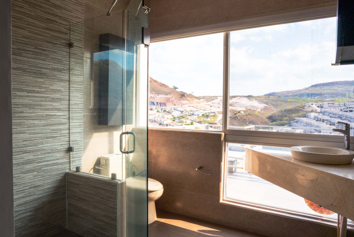 Baño con hermosa vista que cuenta con amplio ventanal que te brindan una hermosa vista, regadera moderna decorada con finos cristales, retrete de color beige y lavamanos de color similar.