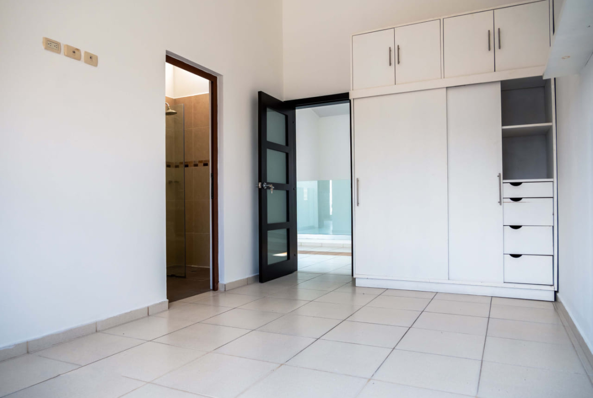 Habitación espaciosa, con paredes de color blanco, suelo de color claro, con closet completo de color blanco, cuenta con baño privado y una puerta de madera oscura decorada con vidrio.