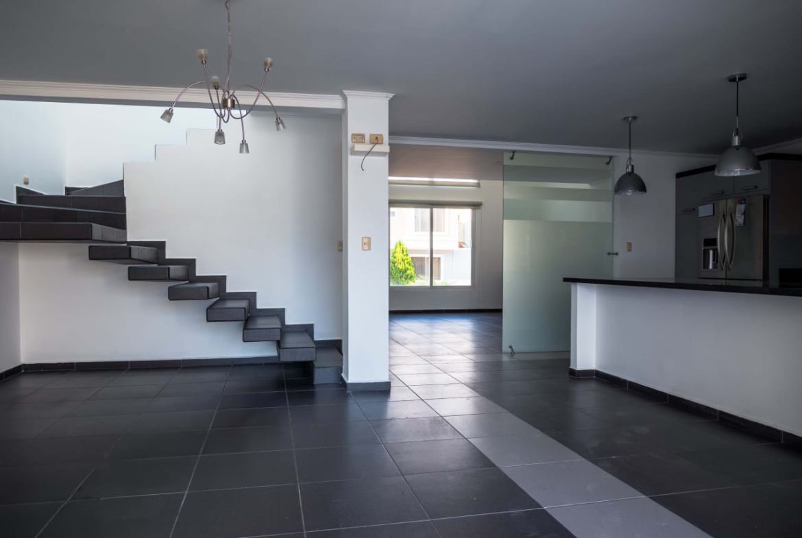 Área de comedor cocina y gradería para el segundo nivel de la casa, las paredes son de color blanco, con suelo de cerámica oscura, adornados con diferentes lamparas colgantes, las escaleras flotantes le dan un toque moderno.