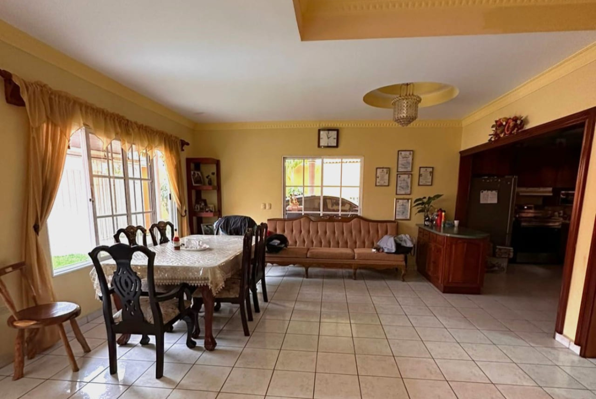 Area de comedor con con paredes de color amarillo y suelo de ceramica blanca, mesa de comedor de madera con 6 sillas de madera oscura, ventanas que dan vista al jardín.
