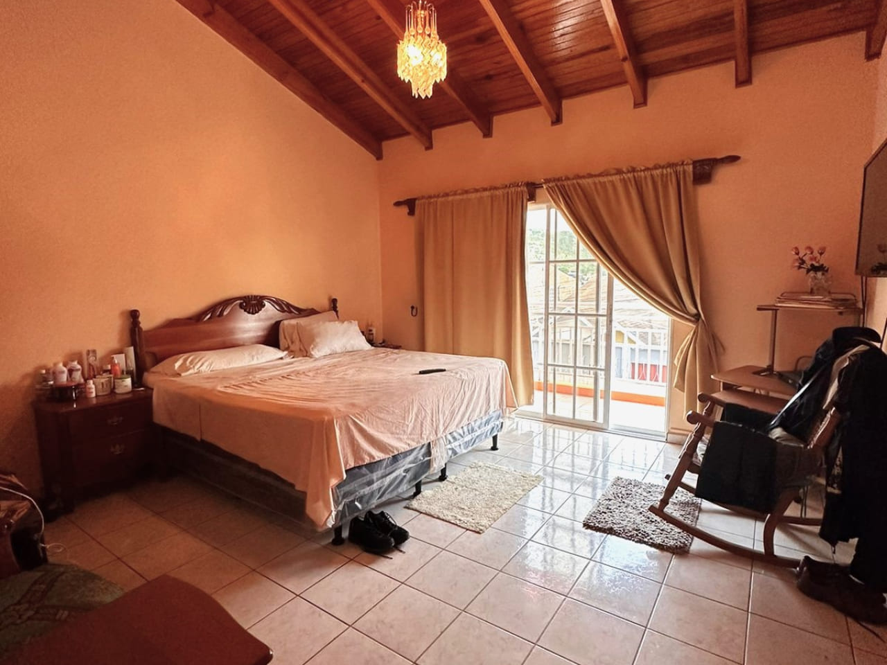 Área de dormitorio con paredes de color carmin, techo de madera, sujujelo de ceramica de color blanco, cama matrimonia, una puerta deslizable que brinda acceso al balcón.