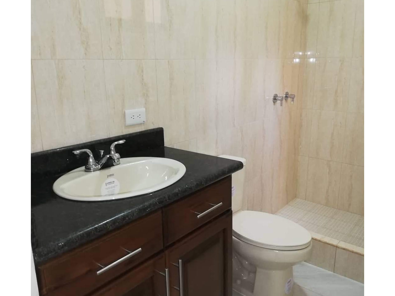 Baño con mueble color café oscuro, plancha color negro en lavamanos junto a inodoro con cerámica en la pared de color blanco.