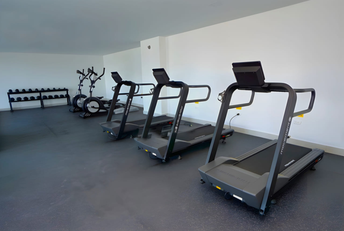 Área de gimnasio con paredes de color claro con diferentes maquinas para realizar cardio y algunas pesas para realizar ejercicios de fuerza.
