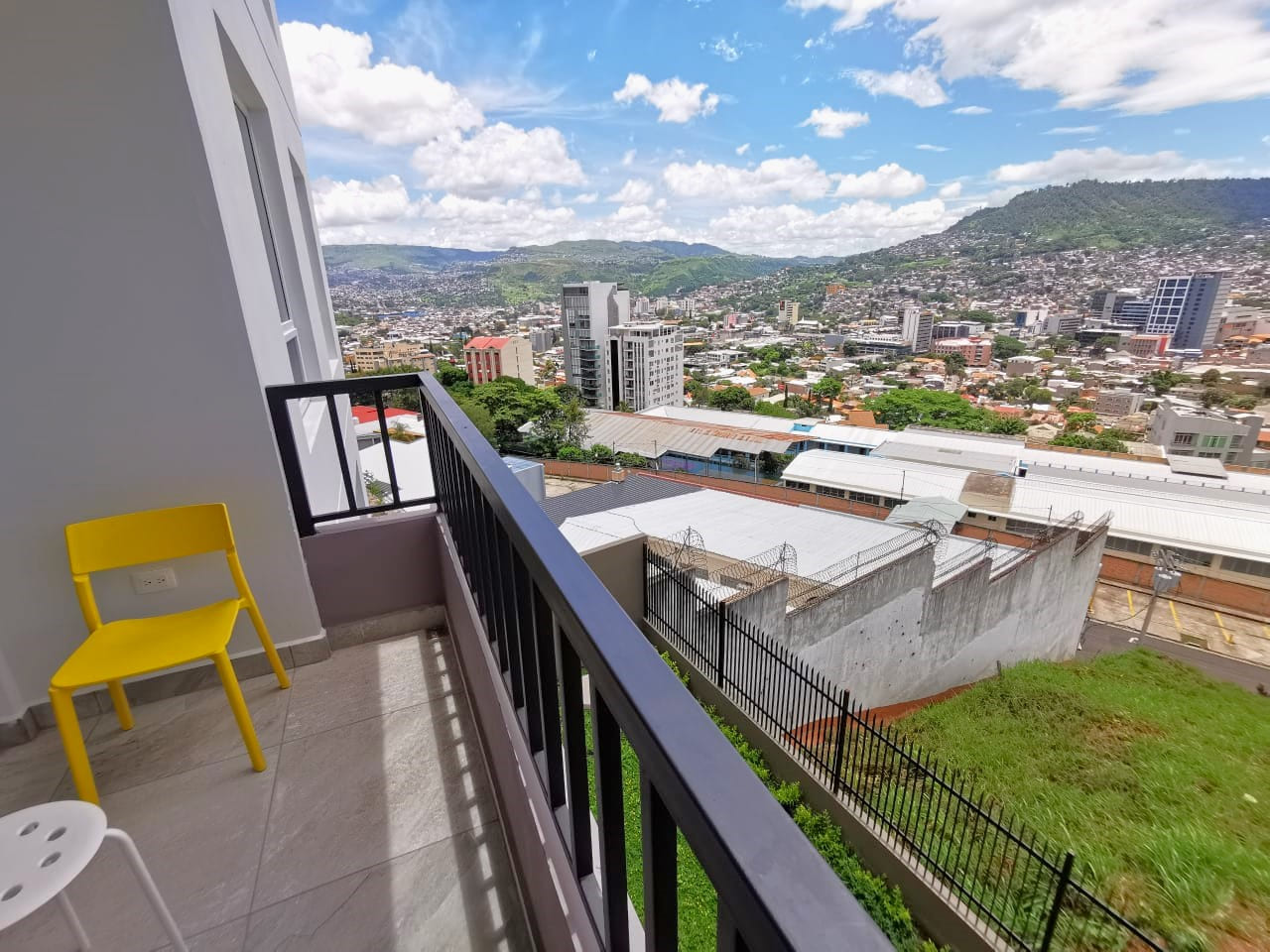 Área de terraza con puerta deslizable, cuenta con un barandal de color oscuro, sillas de color amarillo para apreciar la hermosa vista de la ciudad.