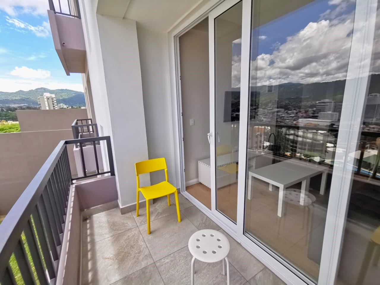 Área de terraza con puerta deslizable, cuenta con un barandal de color oscuro, sillas de color amarillo para apreciar la hermosa vista de la ciudad.