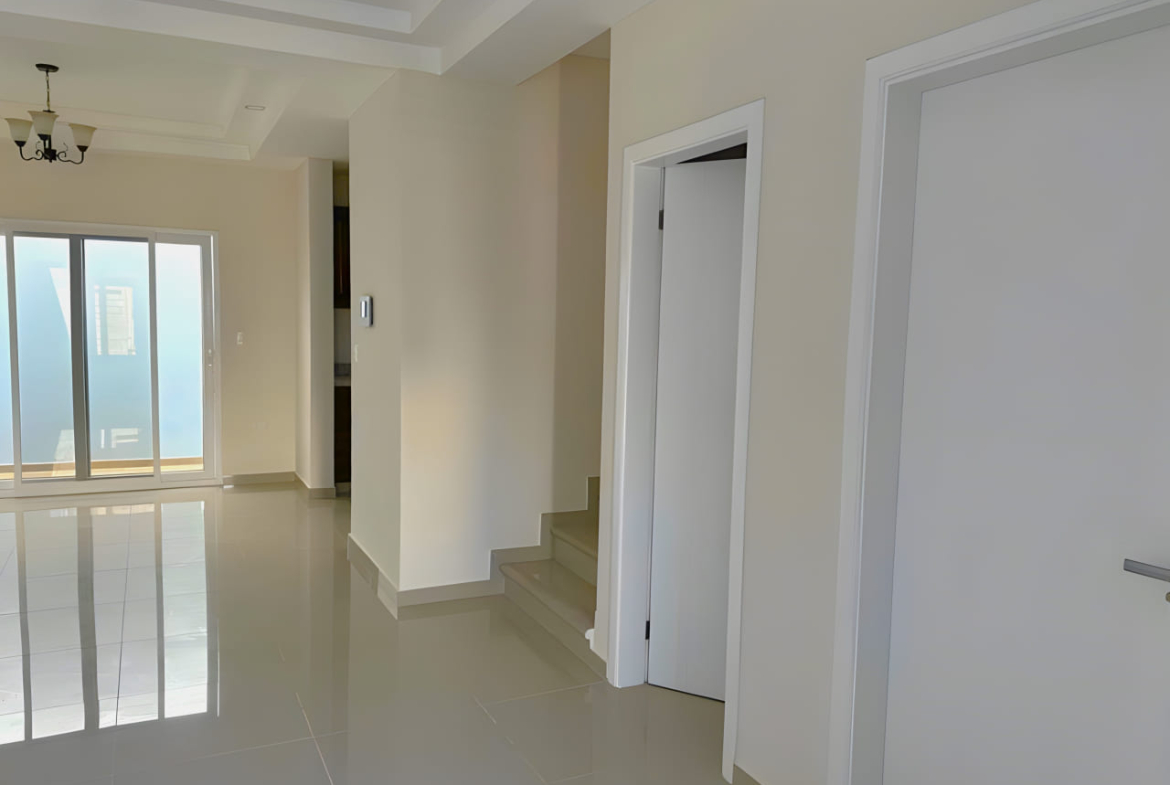 Área de acceso a dos habitaciones y cocina, con puertas de color blanco, suelo de porcelanta, puerta corrediza al fondo que brinda acceso al eterior de la casa.