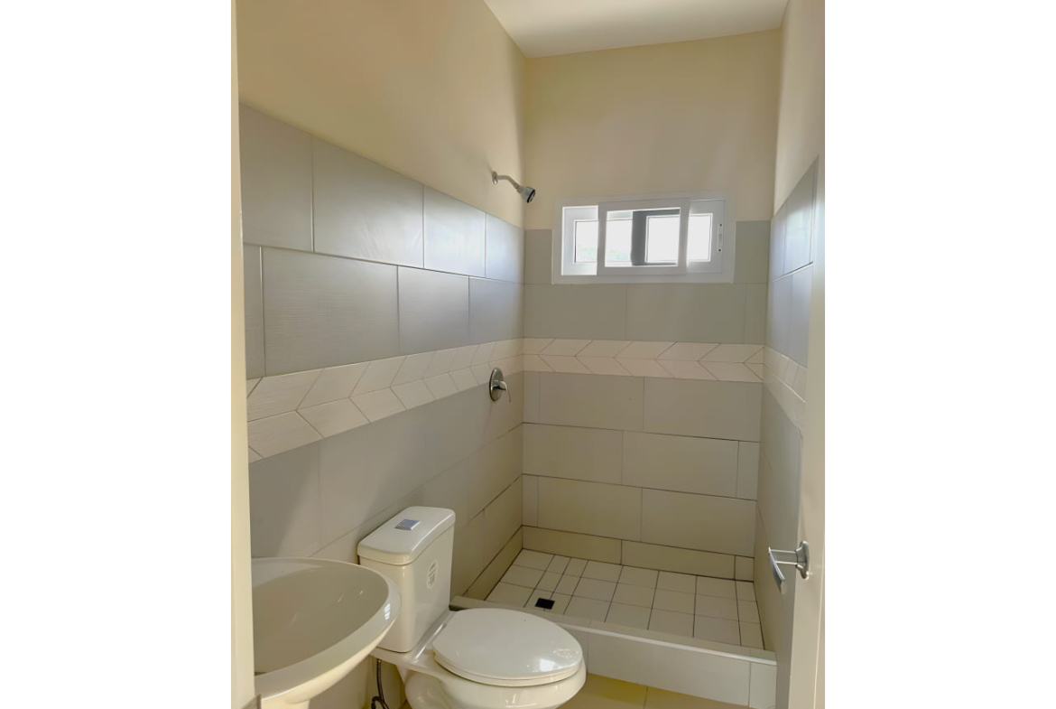 Área de baño cuenta con regadera con suelo de ceramica, retrete y lavamanos de color blanco a juego, con paredes de coradas con ceramica.