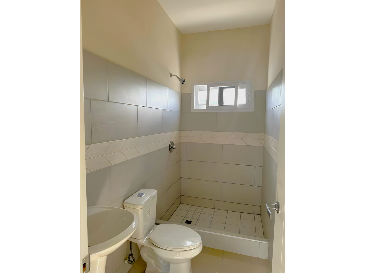 Área de baño cuenta con regadera con suelo de ceramica, retrete y lavamanos de color blanco a juego, con paredes de coradas con ceramica.
