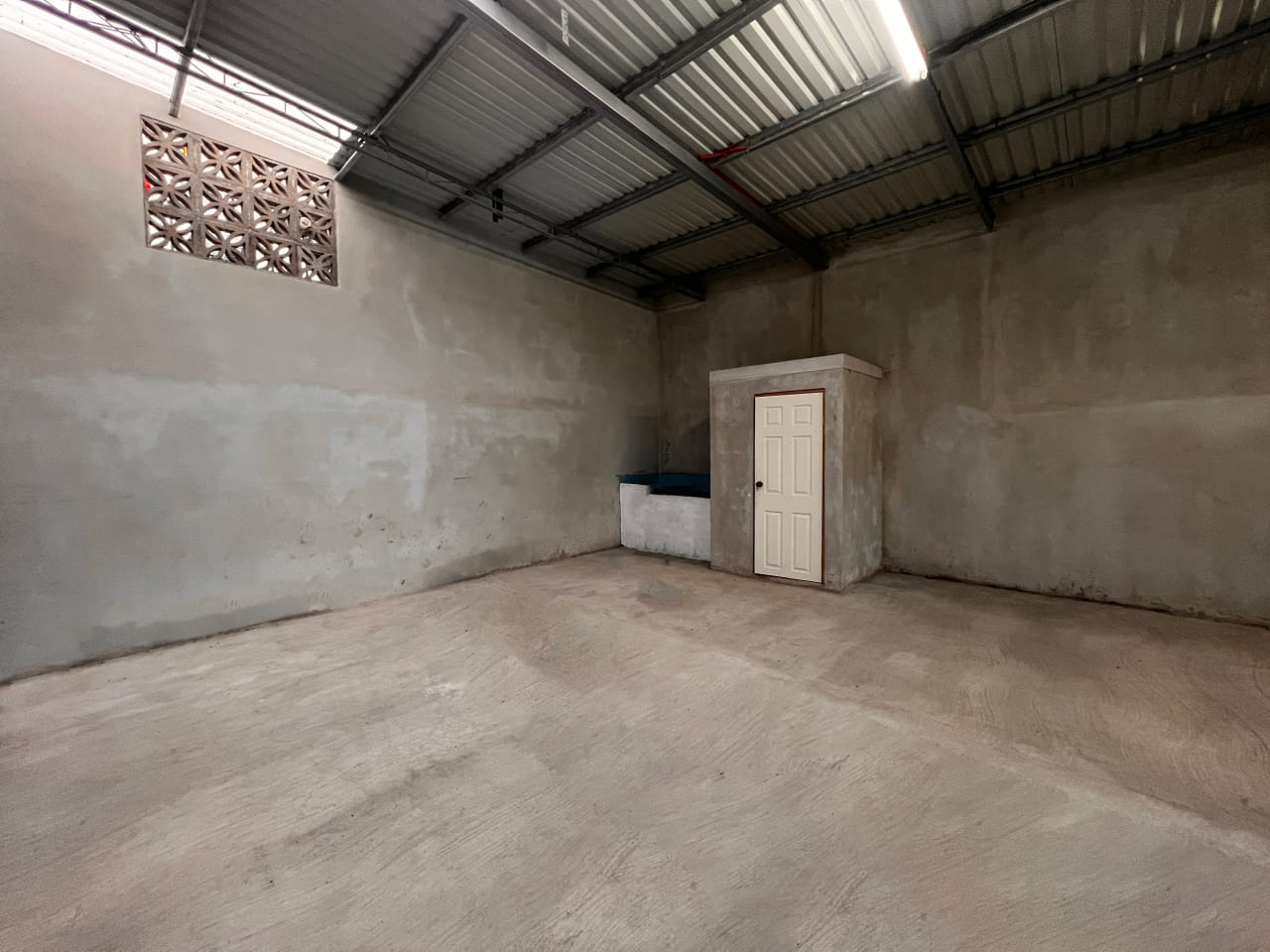 Bodega interior con paredes de bloque, techo de lámina con traga luz, pila para almacenar agua y baño privado al fondo.