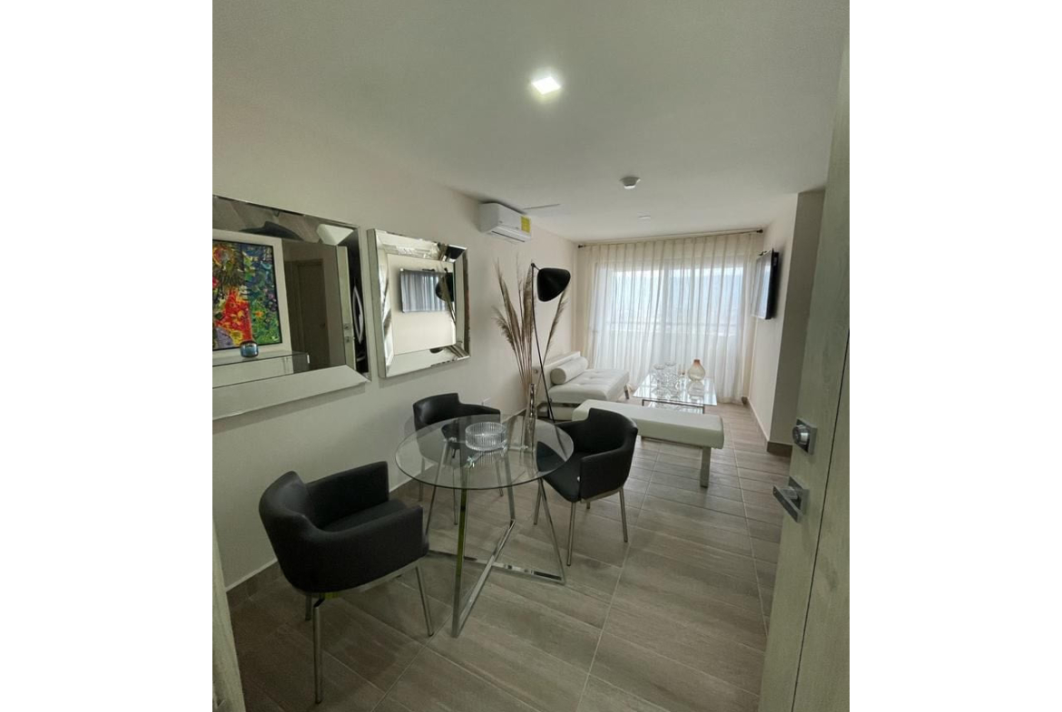 area de sala completamente amueblada cuenta con sillas de color blanco moderno con una mesa de vidrio con accesorios, un televisor colgando en la pared, puerta corrediza que brinda acceso al balcon que cuenta con dos sillas de color negro.