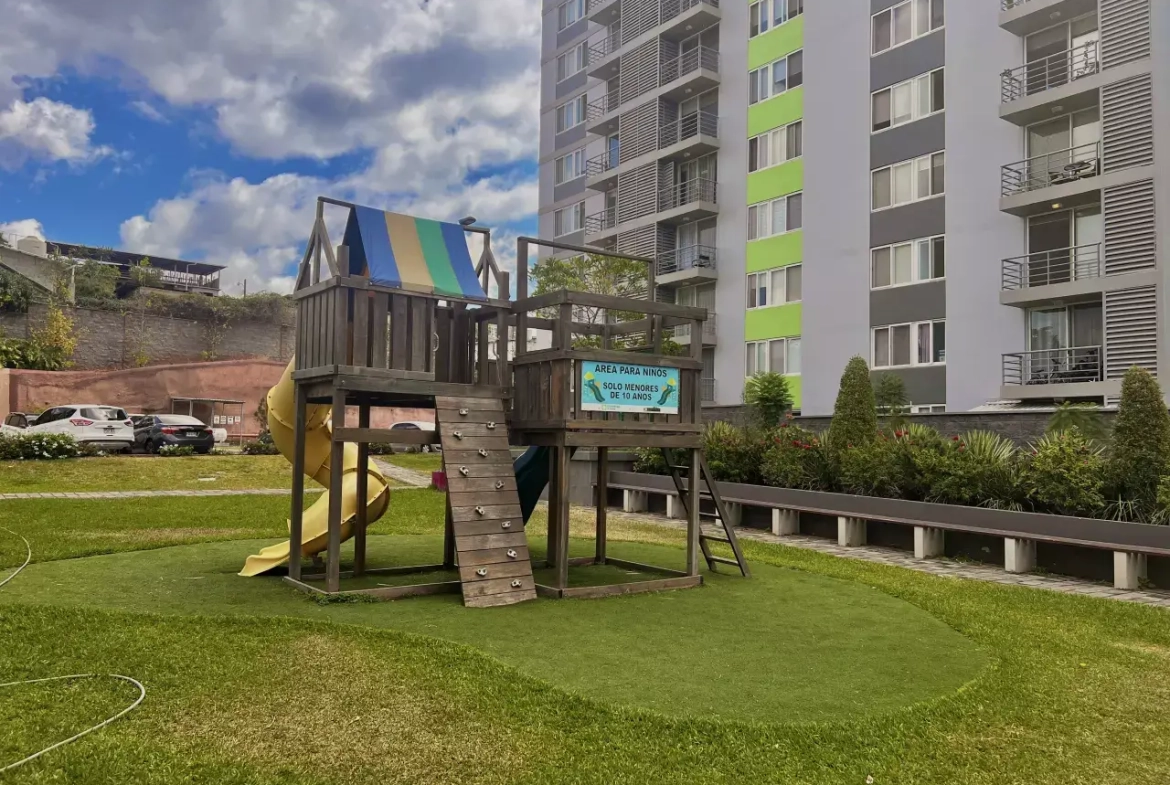 Área de juegos para niños cuentan con una casia de madera que cuentan con una resbaladillas de color amarillo con varia area verde, ademas los condominios cuentan con centro comercial, area de piscina.