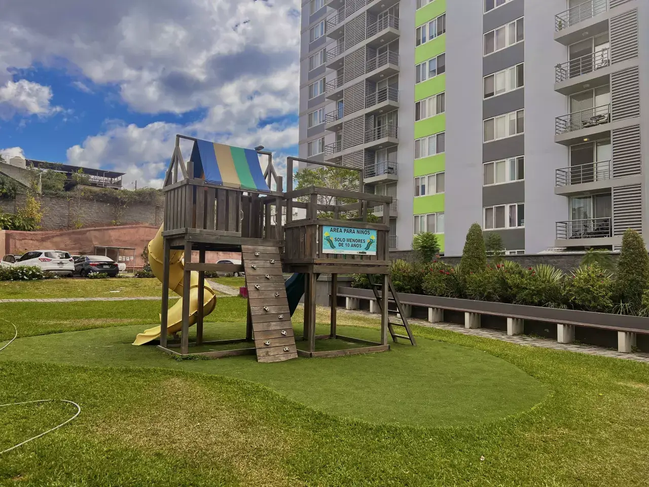 Área de juegos para niños cuentan con una casia de madera que cuentan con una resbaladillas de color amarillo con varia area verde, ademas los condominios cuentan con centro comercial, area de piscina.